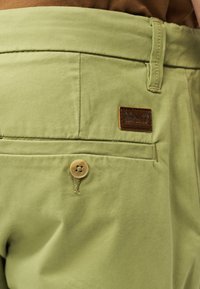 Ανοιχτό πράσινο παντελόνι με πίσω τσέπη που κλείνει με κουμπί και μικρή καφέ ετικέτα με το λογότυπο Timberland πάνω από την τσέπη.