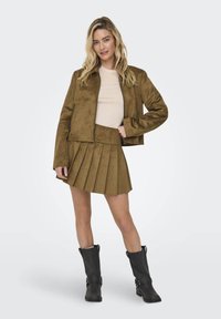Veste cropped en suède marron avec fermeture éclair, associée à une jupe plissée en suède marron et des bottines noires à boucle.