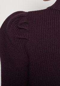 Tmavě fialový žebrovaný svetr s vysokým límcem a nadýchanými rameny, vystavující texturovanou látku a jemné detaily.