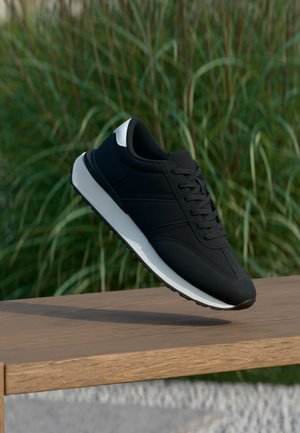 Černá teniska s bílou podrážkou a detailem na patě se vznáší nad dřevěnou lavičkou, zaostřená zelená vysoká tráva v pozadí.