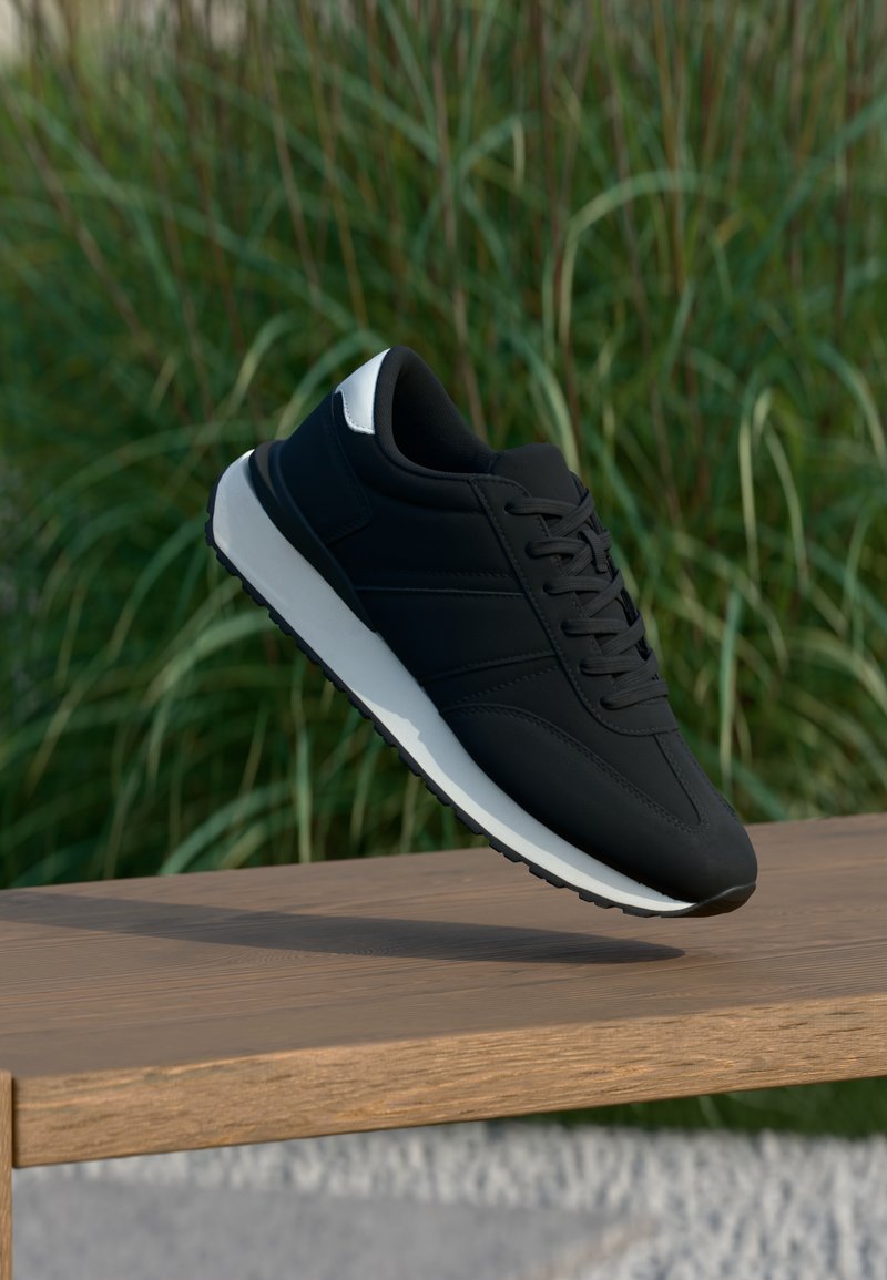 Μαύρο αθλητικό παπούτσι με λευκή σόλα και λεπτομέρεια στη φτέρνα αιωρείται πάνω από ένα ξύλινο παγκάκι, με πράσινο ψηλό γρασίδι θαμπό στο φόντο.