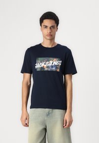 Joven con camiseta azul marino con gráfico floral y texto "Jack & Jones", combinada con jeans de mezclilla clara, posando contra un fondo blanco.