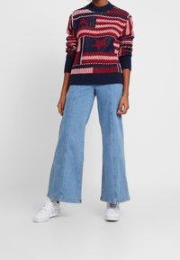 Persoon in een rood, wit en blauw sterrenpatroon trui, met wijde lichtblauwe jeans en witte sneakers, staand tegen een effen achtergrond.