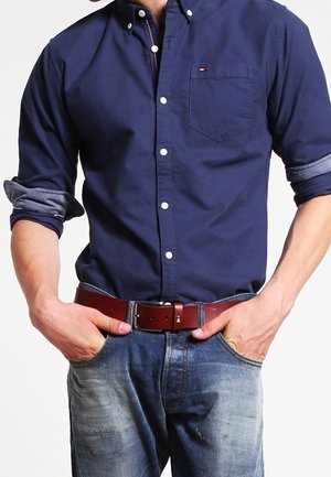 Mężczyzna w granatowej koszuli na guziki z podwiniętymi rękawami, włożonej do niebieskich dżinsów z brązowym skórzanym paskiem, z rękami w kieszeniach.
