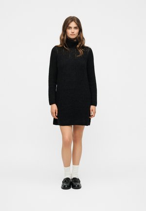 Femme debout portant une robe pull noire à col roulé, des chaussettes blanches et des mocassins noirs sur un fond clair uni.