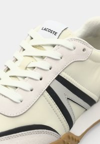 Lacoste tenisice imaju gornji dio od krem sintetike s sivim i crnim detaljima, bijele vezice i teksturiranu gumu na potplatu.