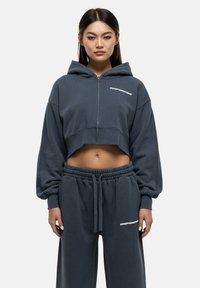 Felpa grigia tagliata con zip frontale, maniche oversize e testo bianco. Abbinata a pantaloni della tuta coordinati con vita elastica.