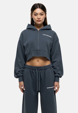 Afgeknipte grijze hoodie met een rits aan de voorkant, oversized mouwen en witte tekst. Gecombineerd met bijpassende sweatpants met een elastische taille.