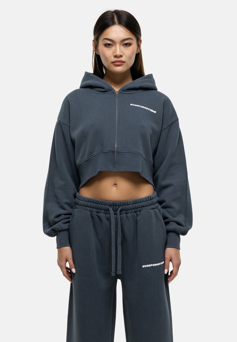 Felpa grigia tagliata con zip frontale, maniche oversize e testo bianco. Abbinata a pantaloni della tuta coordinati con vita elastica.