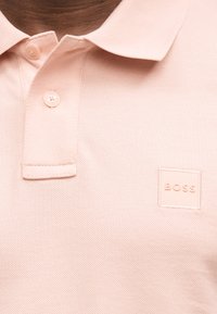 Detail osoby nosiacej svetloružové polo tričko s dvoma gombíkmi a štvorcovým logom "BOSS" na hrudi.