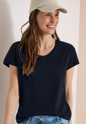 Femme aux longs cheveux bruns portant une casquette beige, un tee-shirt bleu marine à manches courtes et un jean bleu clair, souriante et regardant sur le côté.