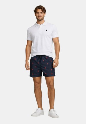Mann trägt weißes Poloshirt mit kurzen Ärmeln und Logo, marineblaue Shorts mit roten gestickten Mustern und weiße Turnschuhe, steht vor einfachem Hintergrund.