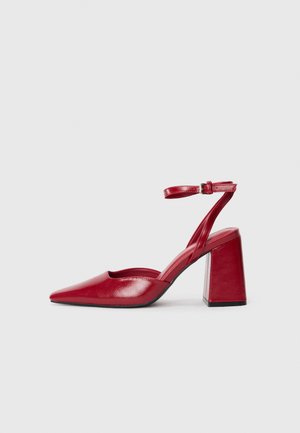 Red leather high-heeled shoes with a pointed toe, open sides, and an ankle strap. Square heel design provides stability. Smooth texture.
