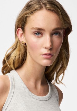 Boucles d'oreilles en or à clous avec un motif floral texturé, portées par une personne aux cheveux ondulés de longueur moyenne, vêtue d'un débardeur gris clair.