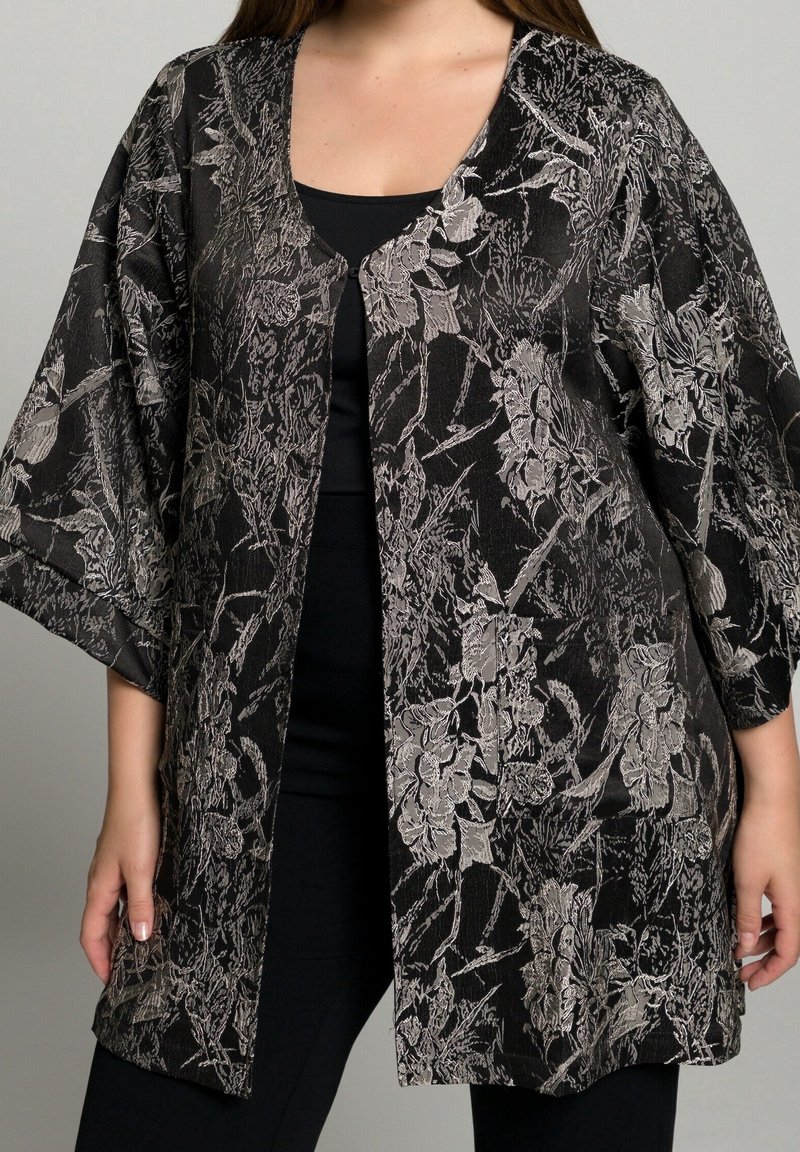 Zwarte kimono-jas met een complex zilverkleurig bloemenpatroon, brede mouwen en een open voorkant. De gestructureerde stof geeft diepte aan het ontwerp.
