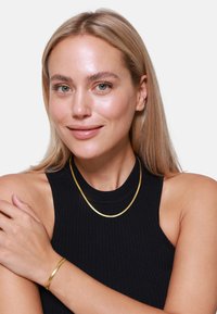 Collana metallica color oro indossata sopra un top senza maniche nero a coste, con una finitura liscia e un design semplice e curvo.