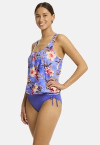 Glunderende vrouw in een blauwe tankini-top met bloemen en een paarse bikinionderkant, zelfverzekerd poserend met één hand op haar heup.