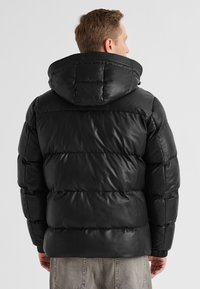 Mann trägt eine schwarze Steppjacke mit Kapuze, von hinten gesehen, kombiniert mit hellgrauen Jeans.