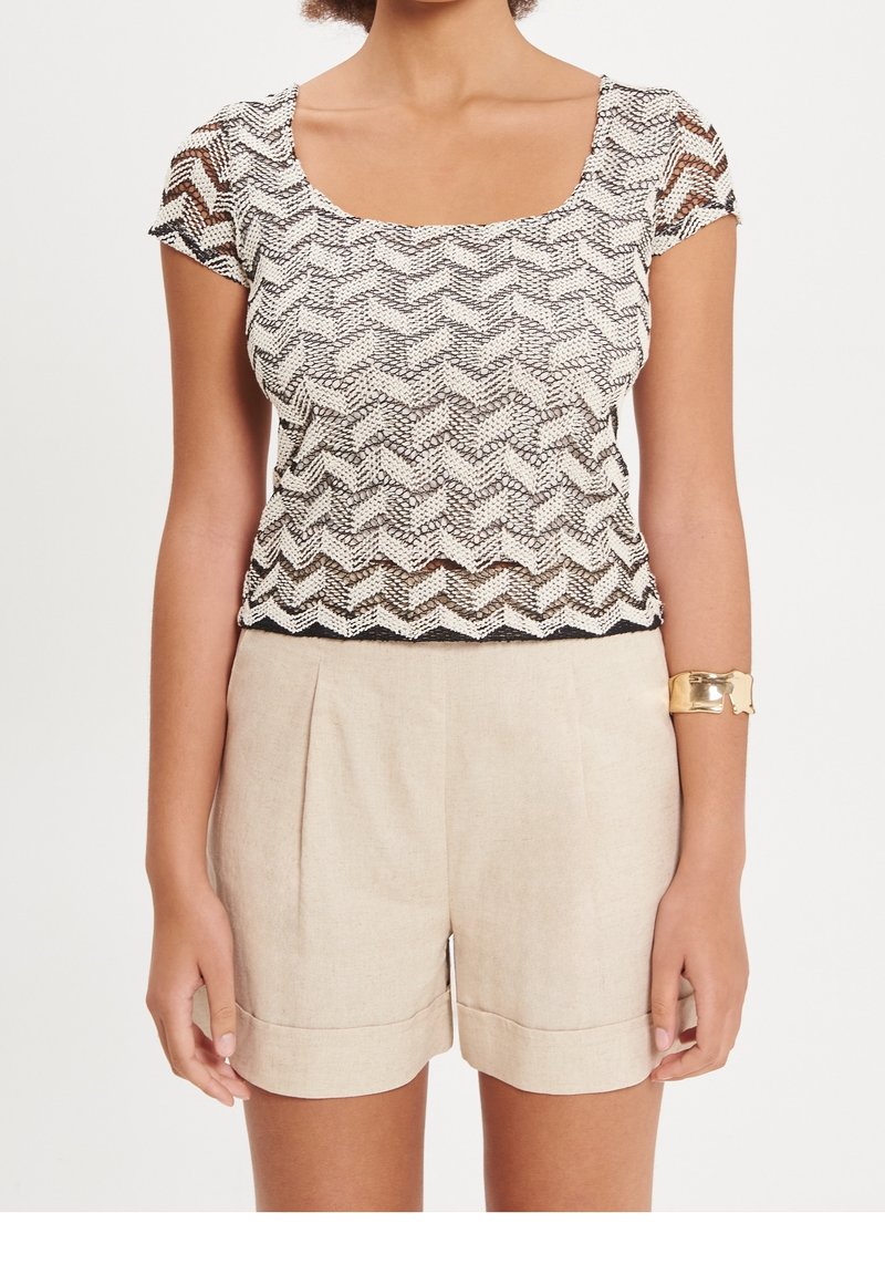 Femme portant un short taille haute beige et un haut en maille à manches courtes crème et noir avec un motif en zigzag, accompagnée d'un bracelet manchette en or.