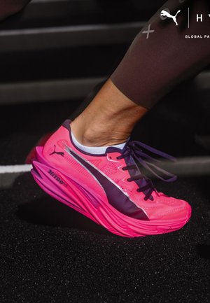 Chaussure de course rose vif avec logo Puma noir et lacets violets, portée par une personne en leggings de sport noirs sur une piste.