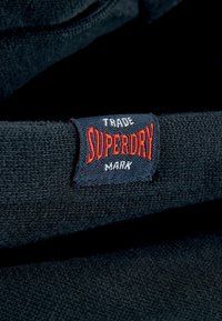 Čierny sveter s modrou a červenou vyšitou nášivkou s nápisom "SUPERDRY." Textúra vyzerá mäkko a pleteno.