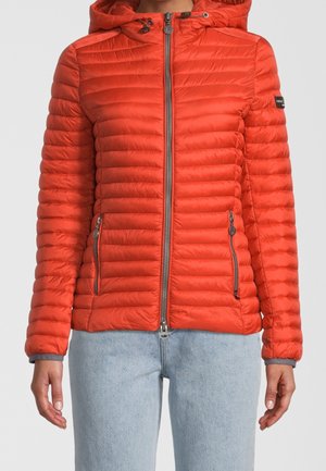 Kobieta nosząca jaskrawo pomarańczową pikowaną kurtkę z kapturem, zamkiem błyskawicznym z przodu i zasuwanymi kieszeniami, w połączeniu z jasnoniebieskimi dżinsami.