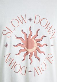 White cotton t-shirt featuring a sun graphic in pink and brown with the text "SLOW DOWN" curved around it, accented by small stars.