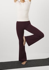 Vaalean beige löysä toppi ja burgundinpunaiset flared-joogahousut, joissa on sivusaumat, seisoo teksturoidulla mustalla matolla, jossa on värikkäitä pilkkuja.