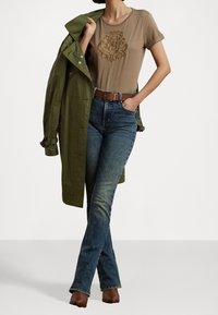 Brun t-shirt med kort ärm och präglad design, i kombination med blå denim flarejeans och en olivgrön trenchcoat, brun bälte och ankelstövlar.