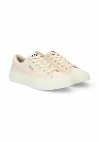 Zapatillas de lona beige con suelas de goma huecas, diseño con cordones, ojales plateados y un logo blanco en la lengua que presenta un patrón de cuadros en blanco y negro.