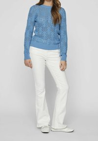 Woman wearing a light blue knitted sweater, white flared pants, and white sneakers standing against a plain background.
