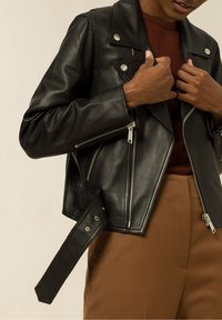 Svart läderbikerjacka med silverdetaljer, fickor med dragkedja och bälte i midjan. Bärs över en vinröd topp och bruna byxor.