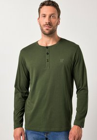 Mies, który ma na sobie długi rękaw, ciemnozielony koszulę typu henley z trzema czarnymi guzikami, zestawioną z niebieskimi dżinsami na jednolitym tle.