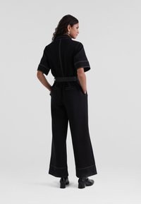 Combinaison noire à manches courtes, col structuré, taille ceinturée, pantalons à jambes larges et couture blanche contrastante. Deux poches à l'arrière.