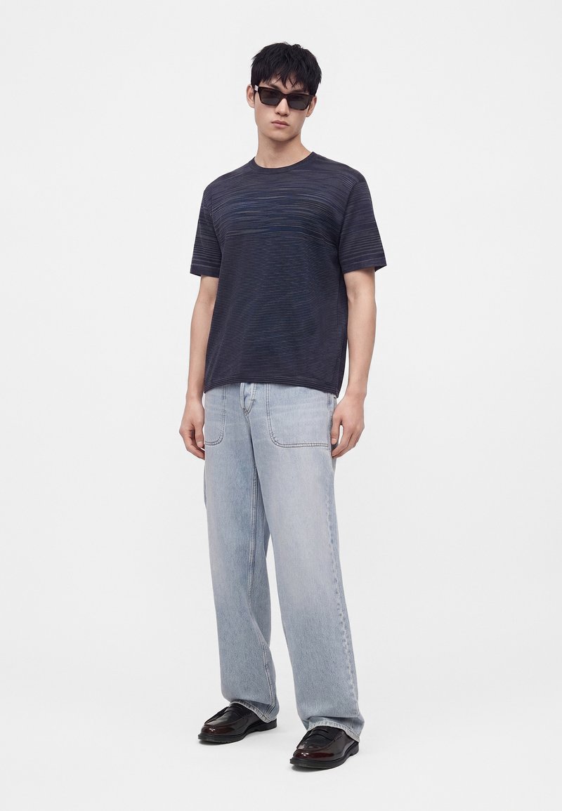 Hombre con camiseta de rayas oscuras, vaqueros de pierna ancha azul claro, zapatos negros y gafas de sol negras, de pie frente a un fondo claro y llano.