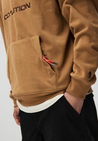 Brązowy polarowy hoodie z kieszenią na przodzie, z czerwonym uchwytem suwaka, ściągaczami oraz haftem "OSTRZEŻENIE" w ciemno-brązowym kolorze.