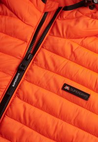 Chaqueta acolchada naranja con costuras horizontales, cremallera negra y una etiqueta con el nombre de la marca "SUPERDRY" en negro sobre un fondo contrastante.
