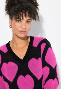 Schwarzer pelziger Pullover mit großen rosa Herzmustern, V-Ausschnitt und geripptem Kragen. Model mit lockigem Haar trägt goldene Creolen.