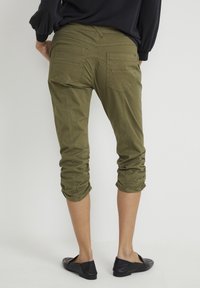 Olive green cropped pants with gathered cuffs, two back pockets, and visible stitching details. Black flats are worn with the pants.