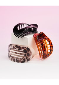 Trois grandes pinces à cheveux en plastique, en noir, écaille de tortue et imprimé léopard, disposées autour d'un cube blanc sur un fond rose.