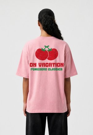 Person med krøllet hår i en hestehale bærer en lyserød skjorte med to røde tomater og teksten "ON VACATION POMODORO CLASSICS" på bagsiden.