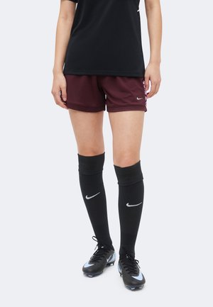 Personne portant un maillot de sport noir, un short bordeaux, des chaussettes Nike noires montant aux genoux et des chaussures de football Nike noires, debout devant un fond uni.