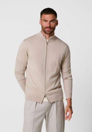 Uomo con capelli corti e scuri e barba, indossa un maglione beige con zip e pantaloni grigio chiaro su uno sfondo chiaro uniforme.