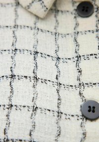 Tweedtyg i krämfärg med ett svart rutmönster; har svarta knappar och en krage med synliga sömnadsdetaljer.