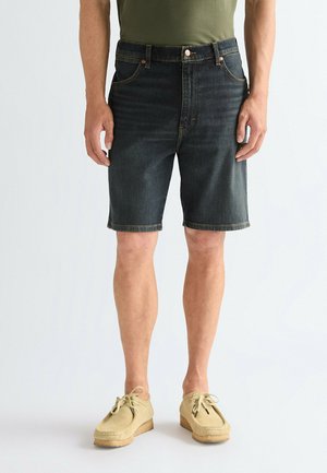 Mann trägt dunkelblaue Jeansshorts, olivgrünes T-Shirt und beige Schnürschuhe, steht vor einem schlichten Hintergrund.