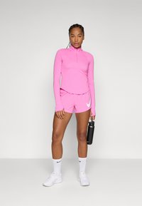 Rózsaszín sport hosszúujjú felső félnyitott cipzárral, hozzáillő rövidnadrág, fehér Nike logóval díszítve, fehér zokni, és egy fekete vízpalackot tart a kezében.