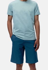 Man i ljusblå kortärmad t-shirt med diskret tryckta bokstäver och mörkblå knälånga shorts, stående med en hand i fickan.