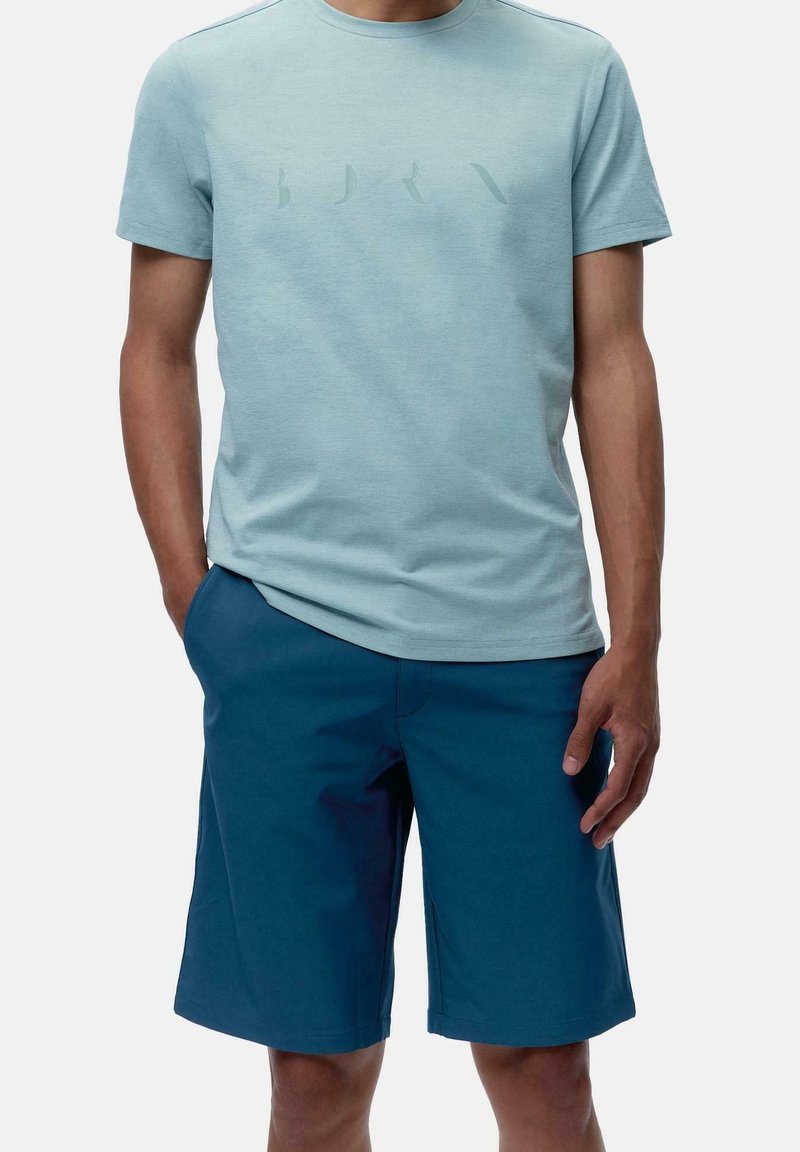 Man i ljusblå kortärmad t-shirt med diskret tryckta bokstäver och mörkblå knälånga shorts, stående med en hand i fickan.