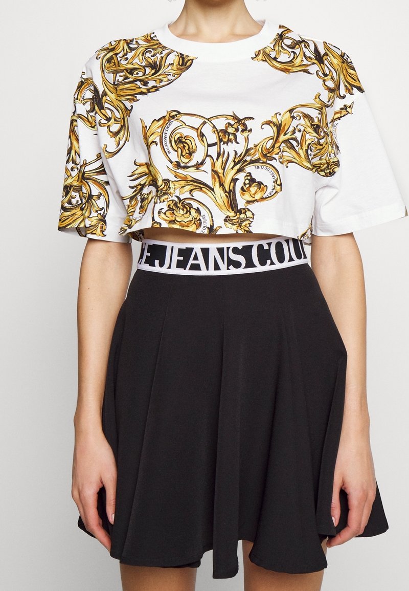 Zkrácený bílý tričko s zlatými a černými květinovými vzory, doplněné černou sukní s kontrastním bílým pasem označeným "JEANS".
