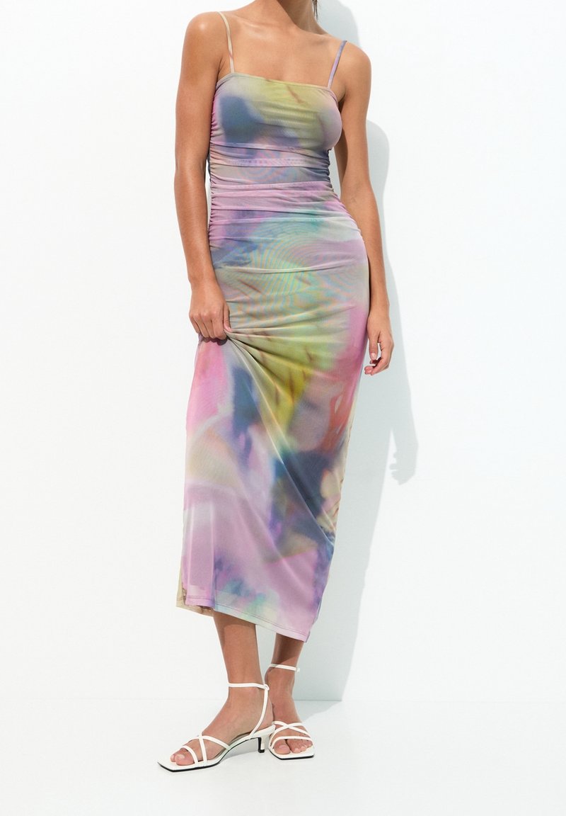 Femme portant une robe midi ajustée sans manches, multicolore avec motif tie-dye, et des sandales blanches à talons avec brides, debout devant un fond blanc.